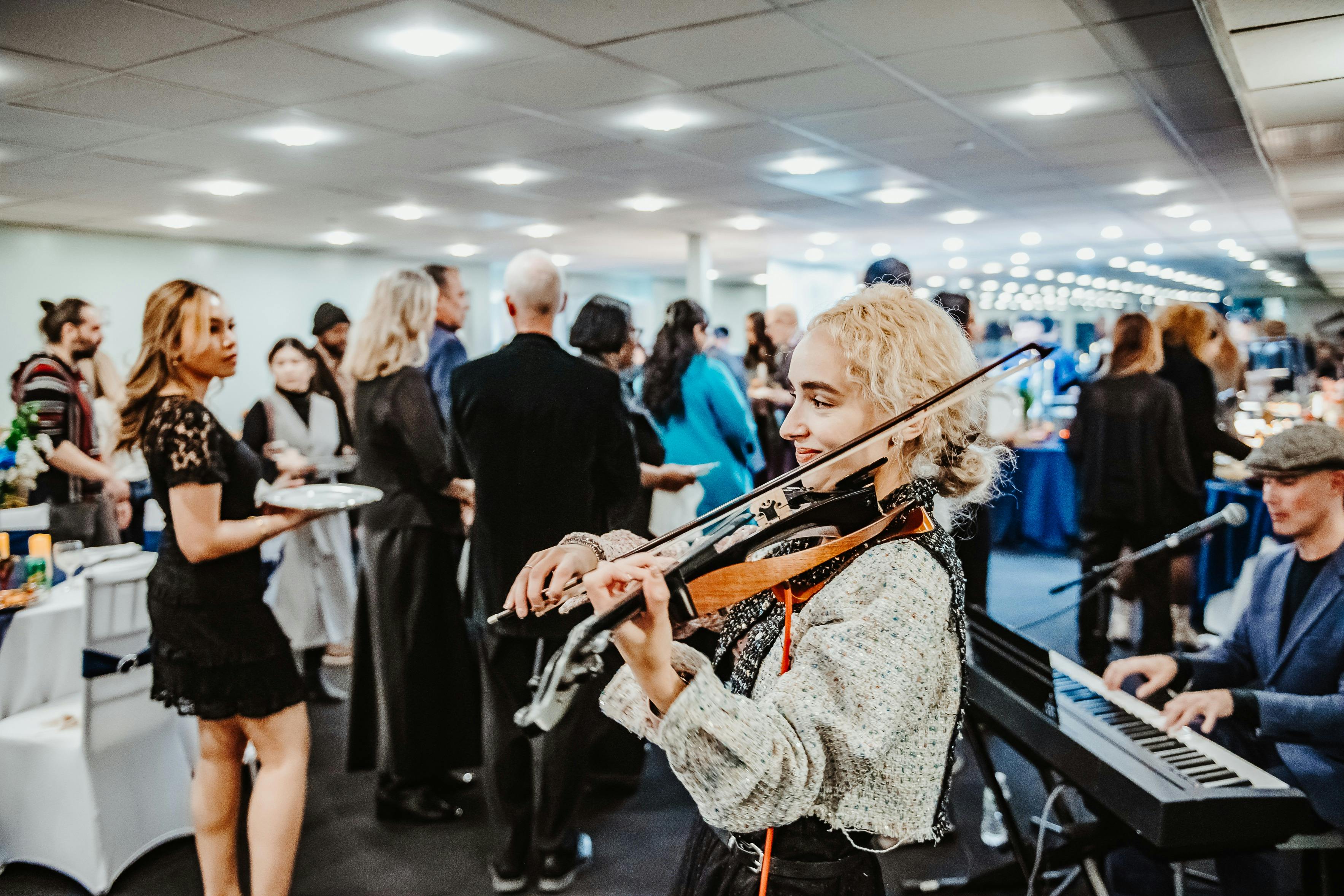 Žena hraje na housle, zatímco muž hraje na klávesy na halové akci s lidmi, kteří se socializují v pozadí.