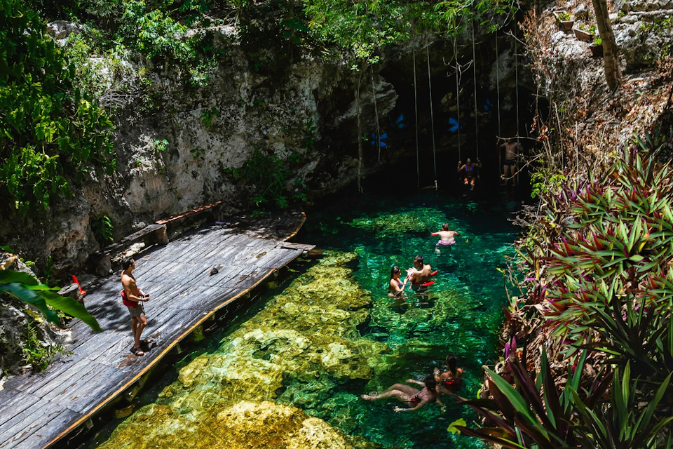 People swim in a clear, turquoise cenote surrounded by rocky walls and lush greenery, with some standing on a nearby wooden deck.