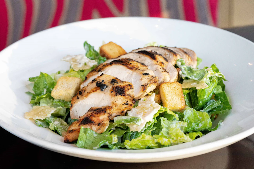 Grilled chicken slices on a Caesar salad with romaine lettuce, croutons, and Parmesan cheese in a white bowl.