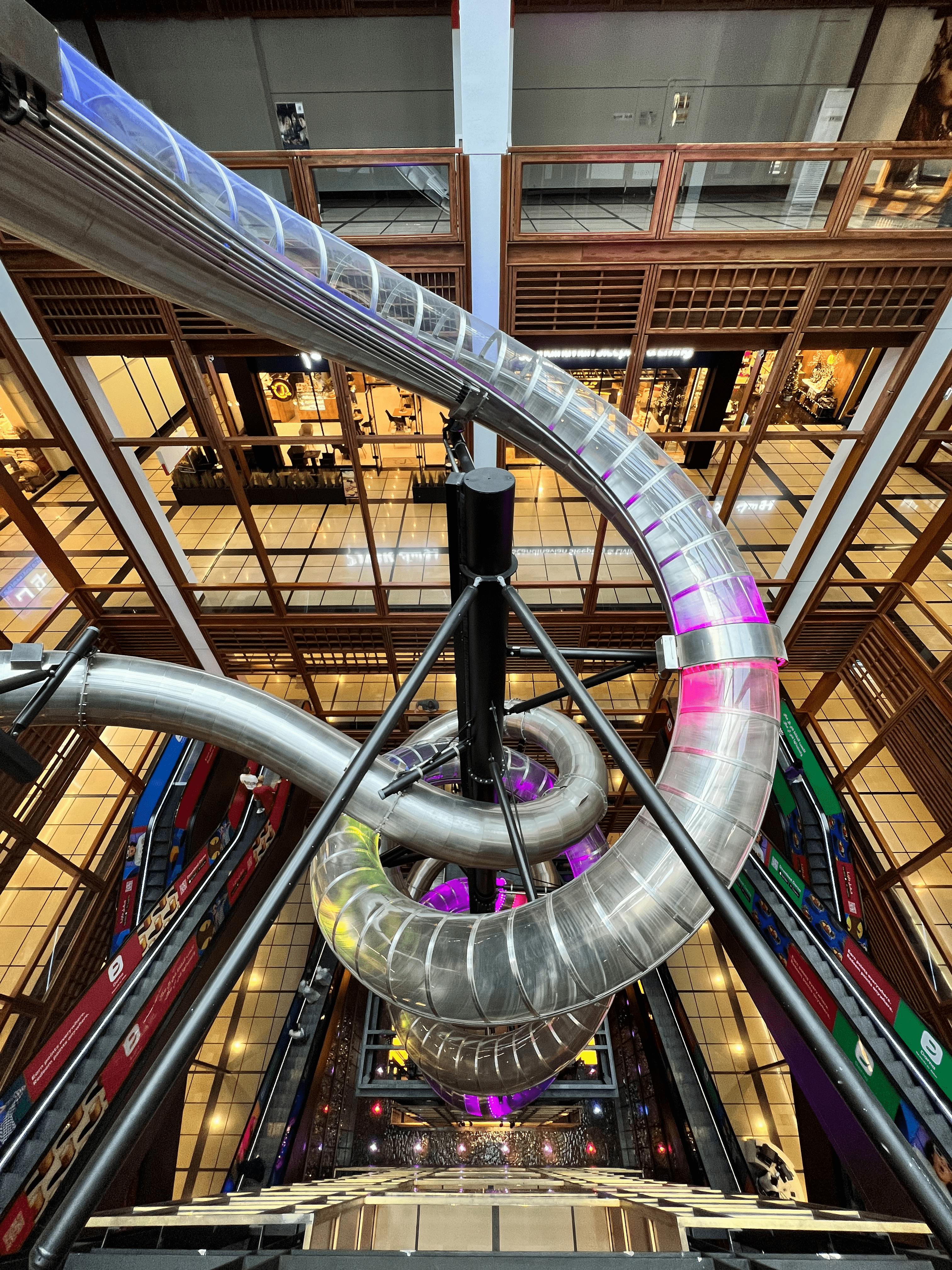 Vysoký, vícepodlažní vnitřní skluzavka s kovovými trubkami spirálovitě dolů, zasazený do budovy s dřevěnými a skleněnými stěnami.