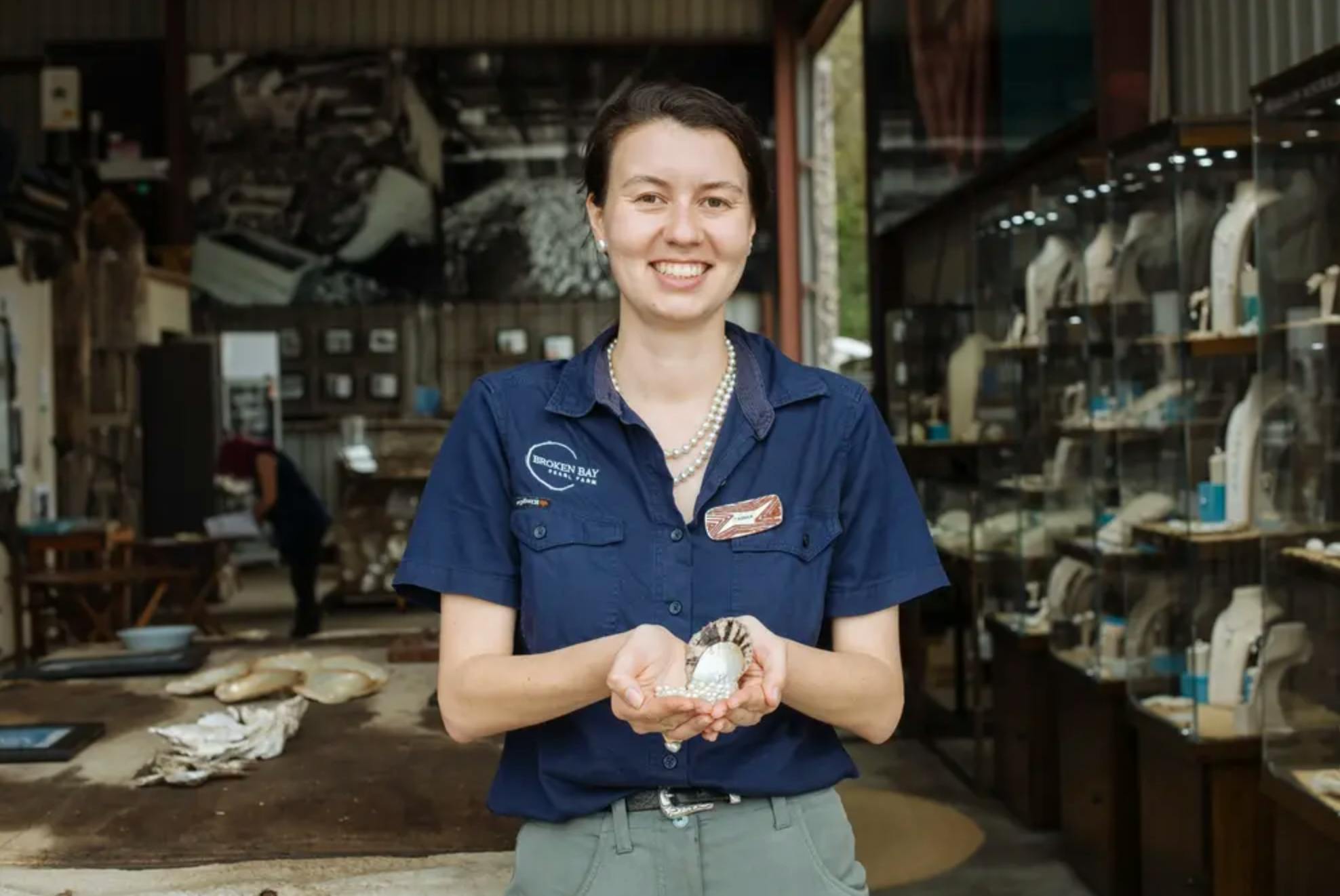 Una persona con una camicia blu con cartellino tiene in mano una conchiglia con una perla all'interno, in un ambiente simile a un laboratorio.