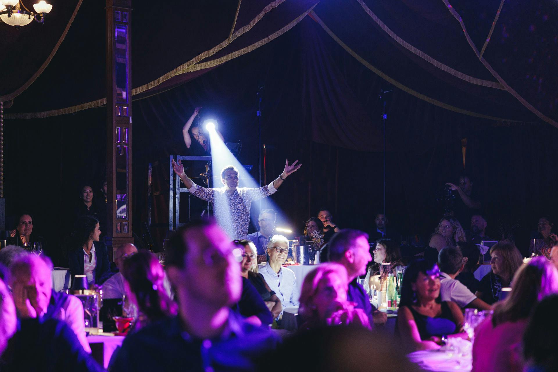 聚光灯照射在一个拥挤、光线昏暗的房间里举起手臂的人身上，房间里有桌子和坐着的房客。