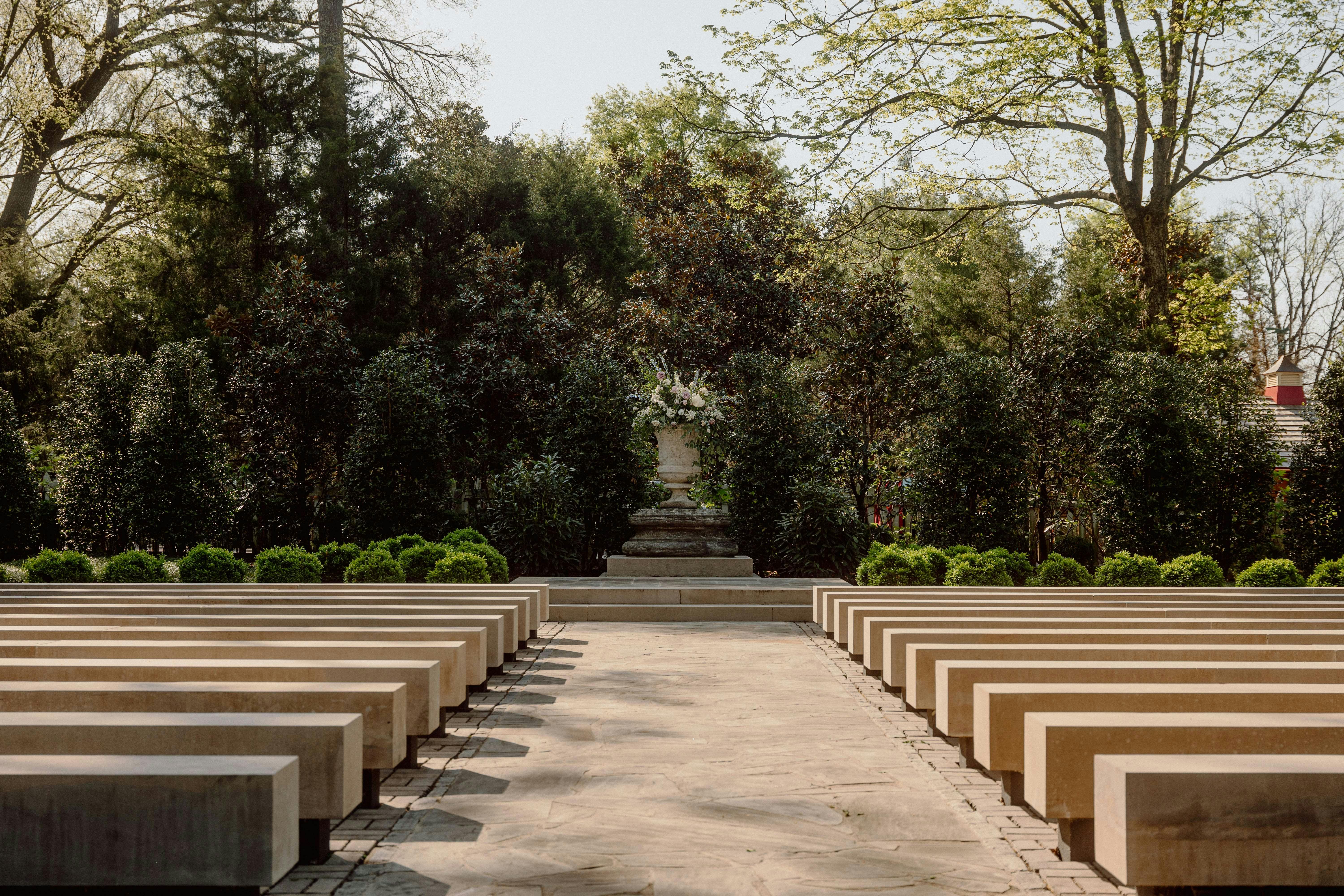 Venkovní ulička s kamennými lavičkami po obou stranách, vedoucí k podstavci s velkým květinovým aranžmá, obklopenému stromy a zelení.