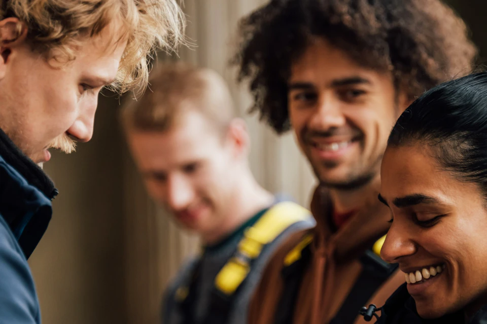 A group of people preparing for an activity, with one person assisting another with safety harnesses, all smiling.