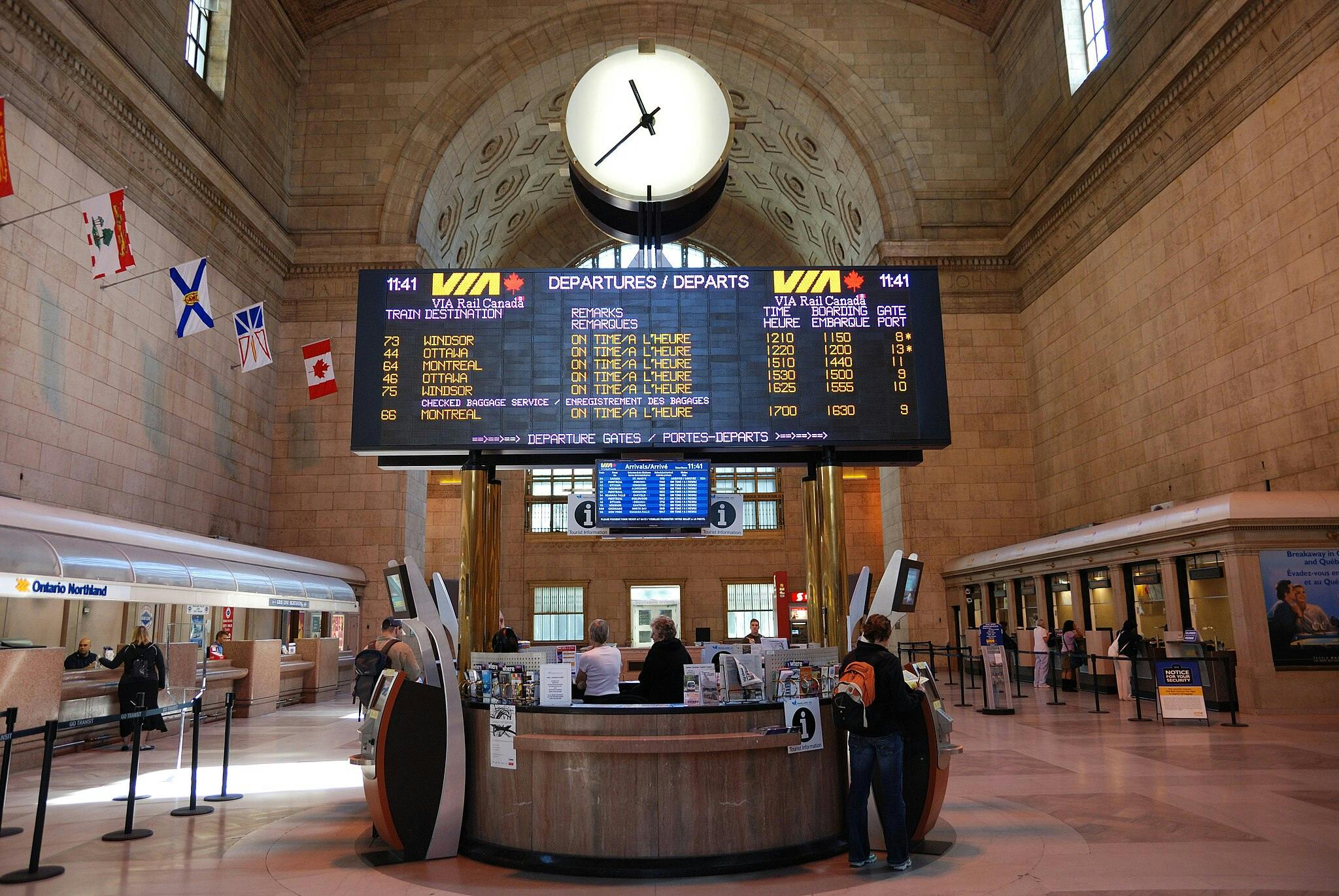 Interior of a grand train station with a large departure board, information center, flags, and people at the ticket counters.