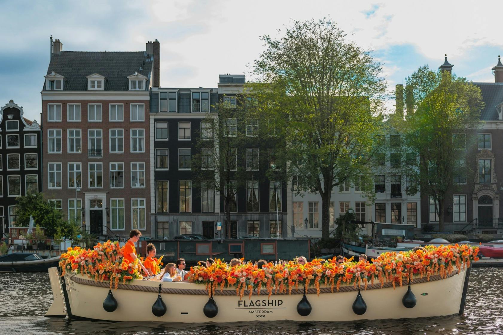 Ein mit Blumen geschmücktes Boot mit Passagieren an Bord, das auf einem Kanal treibt, im Hintergrund sind historische Gebäude und Bäume zu sehen.