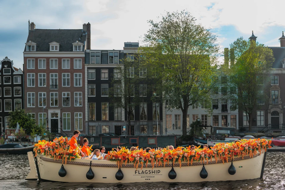A boat decorated with flowers and carrying people, floating on a canal with historic buildings and trees in the background.