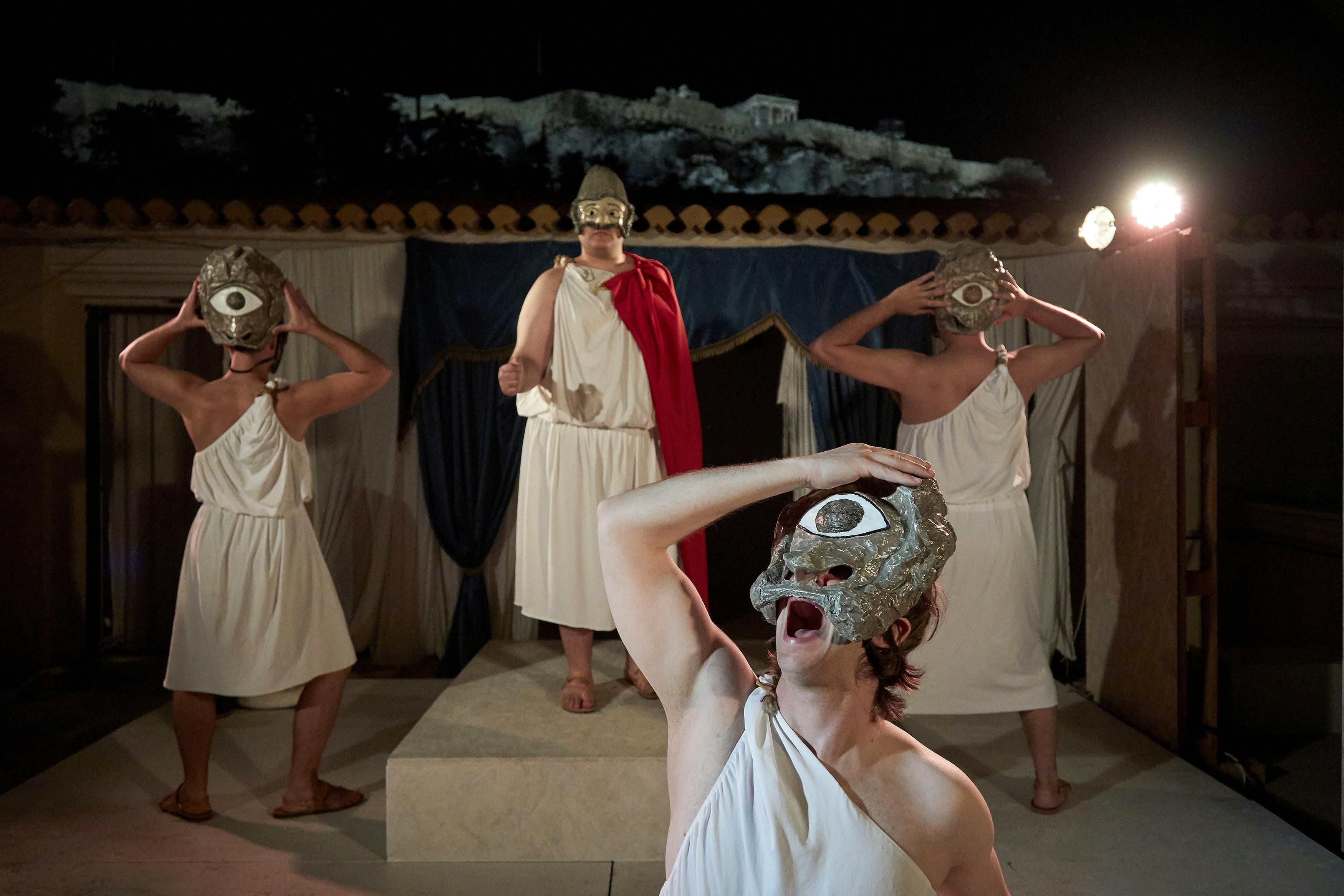 四个戴着白色长袍和眼罩的人在一个有窗帘和石头废墟背景的舞台上表演古希腊戏剧。