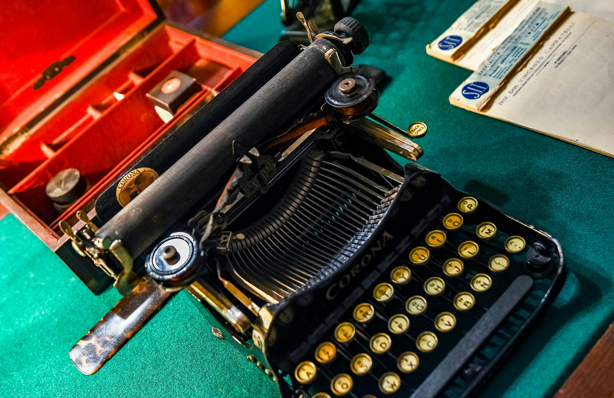 En vintage Corona skrivemaskine med gule taster på en grøn overflade, ved siden af en rød kasse og nogle dokumenter.