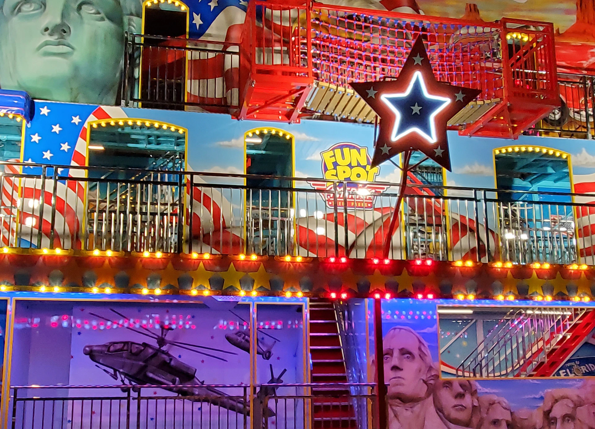 A brightly lit amusement attraction themed around America, featuring the Statue of Liberty, Mount Rushmore, and patriotic decorations.