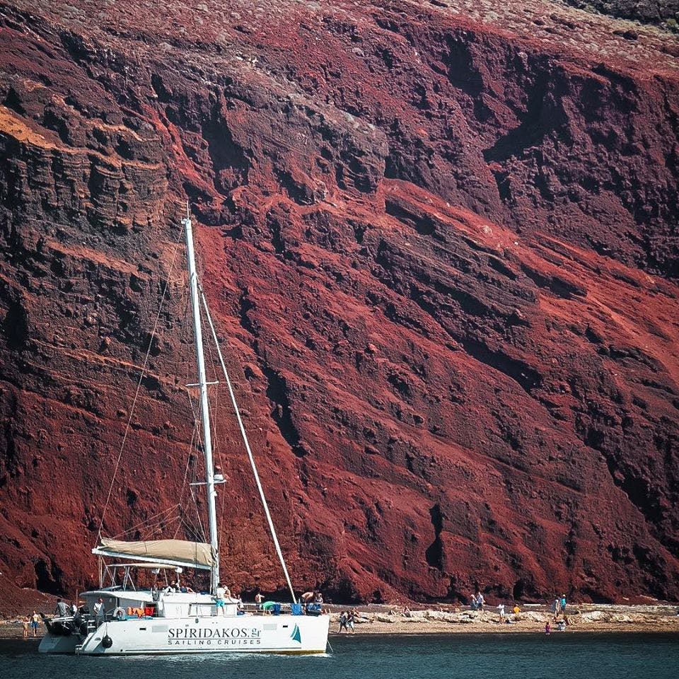 험준한 붉은 바위 절벽 근처에는 흰 범선이 떠 있고, 해변을 배경으로 사람들이 거닐고 있습니다.