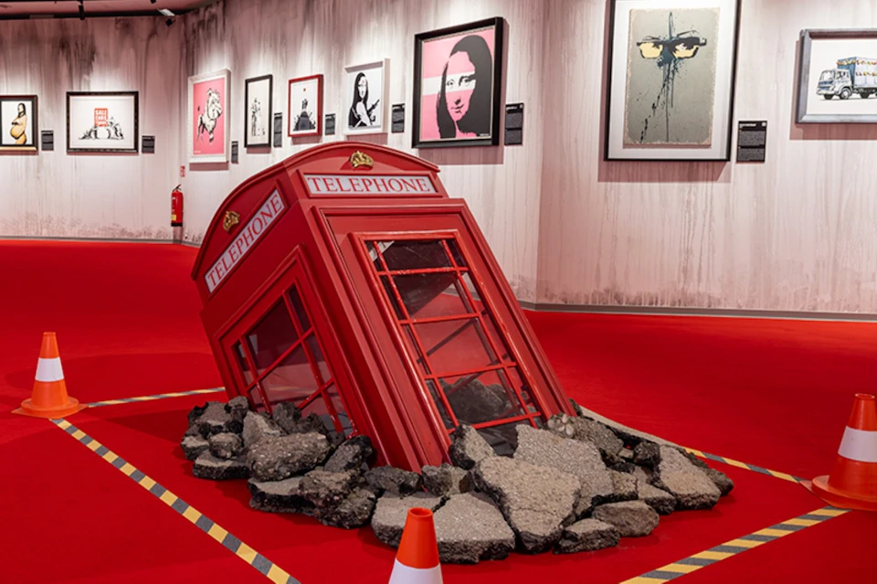 A red British telephone box partially sunken into rubble in an exhibition space with framed artworks on the walls.