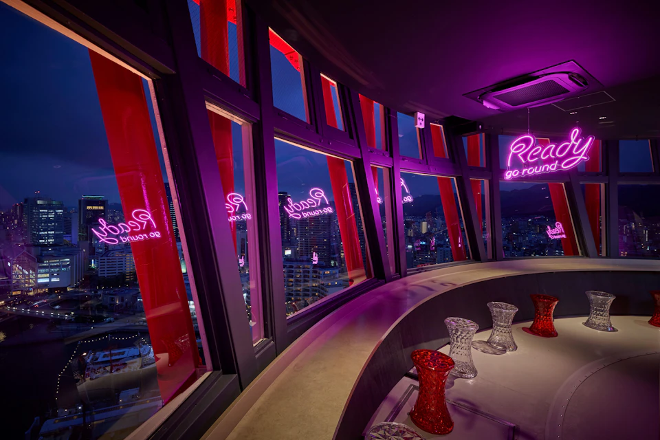 A modern lounge with curved seating, red and clear glassware on the table, and large windows displaying a city view at dusk.
