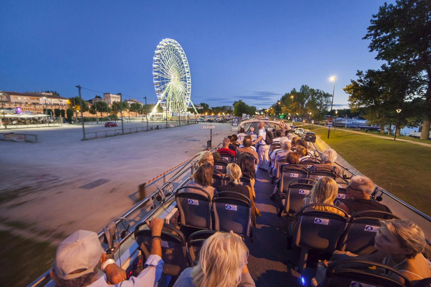 黄昏时分，人们坐在一辆敞篷巴士上，朝着一个明亮的摩天轮走去。 还可以看到树木和路灯。