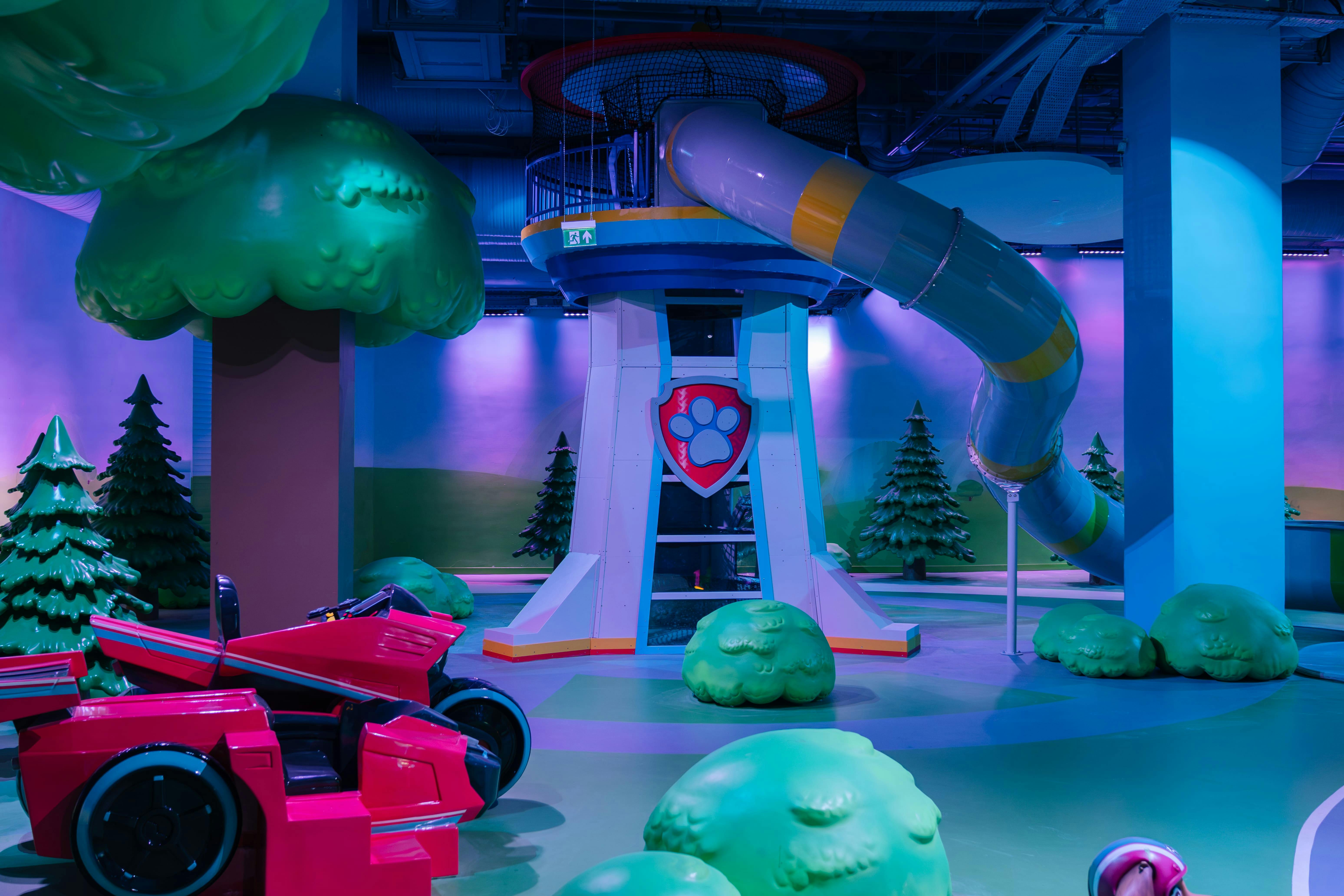 Binnenspeeltuin met bomen als thema, een toren met glijbaan en een rood voertuig, verlicht met kleurrijke lampjes.