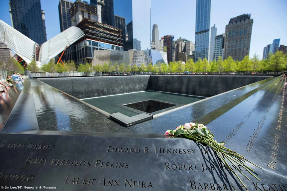 Memorial water fountain with names inscribed, surrounded by skyscrapers. Pink and white flowers are placed on the edge.