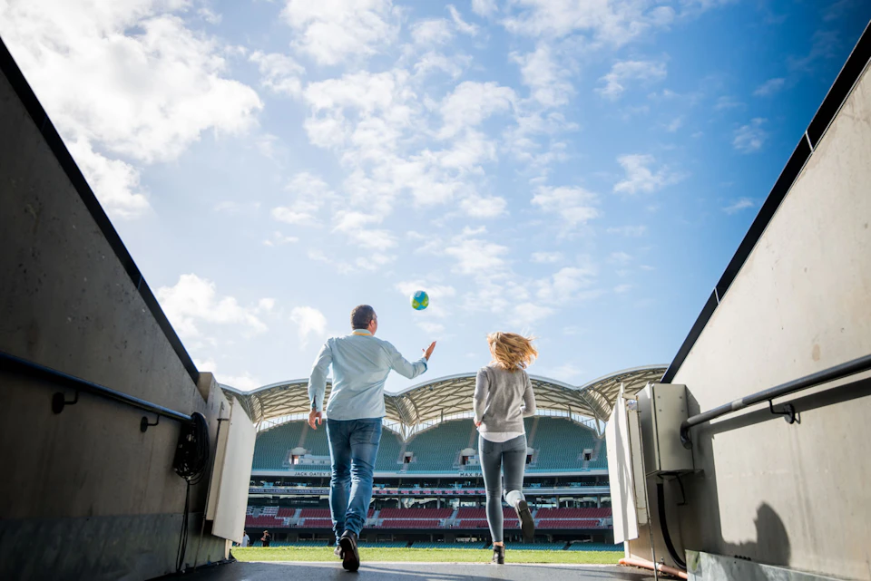 Two people walk towards a sports stadium field under a blue sky, with one person holding a ball.