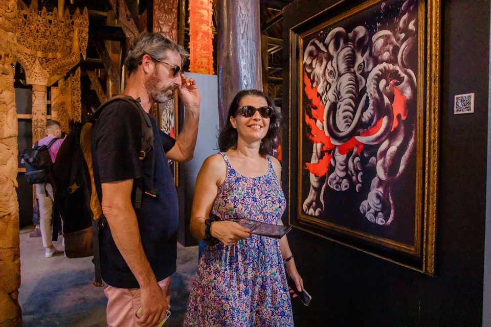 Two people wearing sunglasses view artwork of an elephant in a gallery with wooden decor and colorful details.