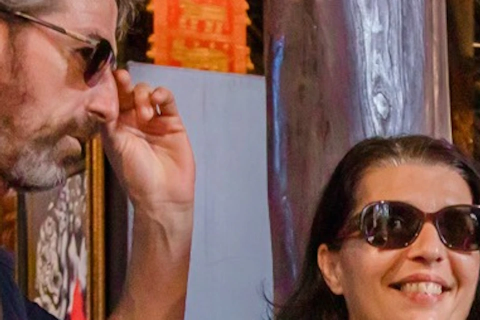 Two people wearing sunglasses view artwork of an elephant in a gallery with wooden decor and colorful details.