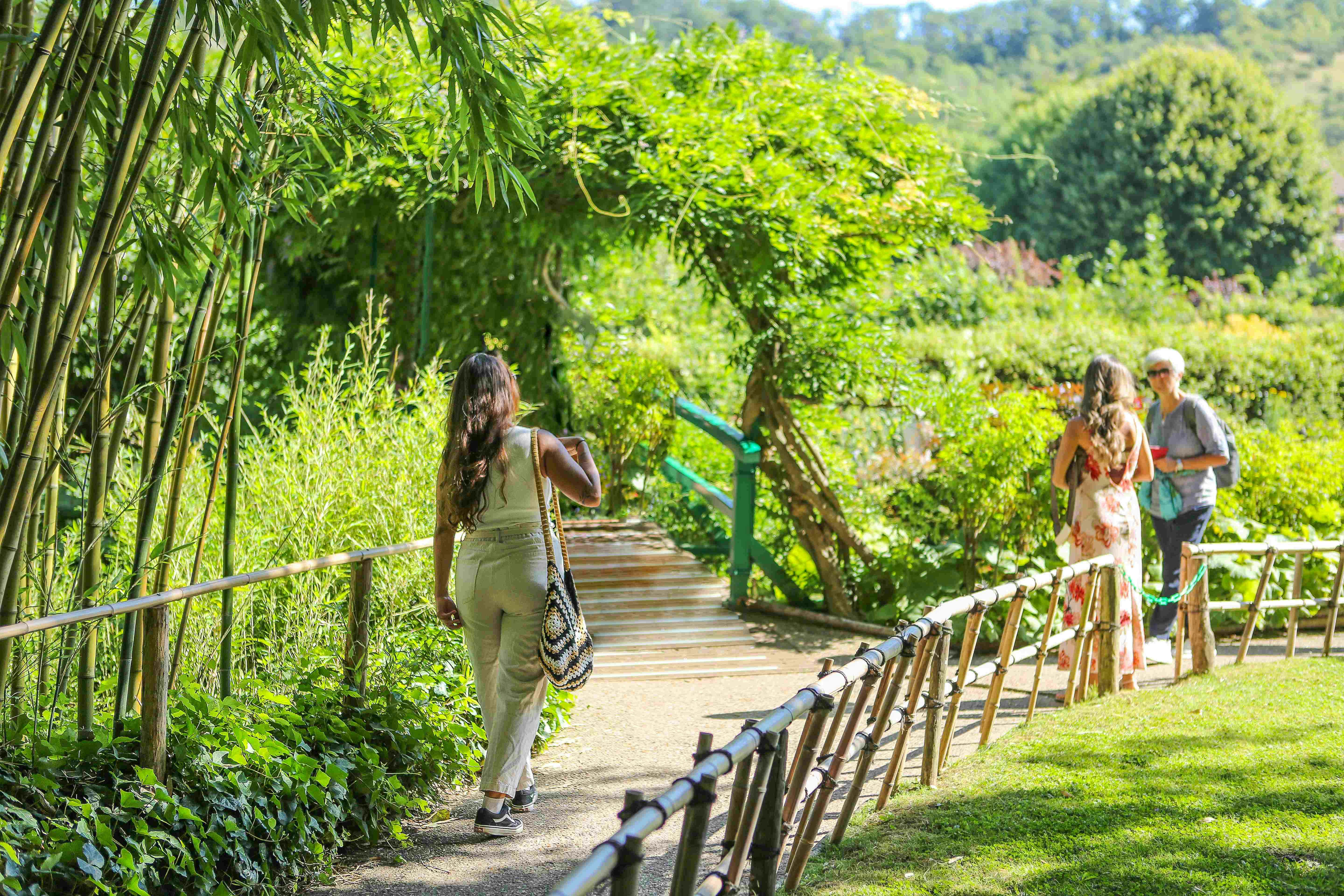 Two women walk along a sunlit garden path lined with lush greenery and a wooden handrail, approaching a vine-covered archway.