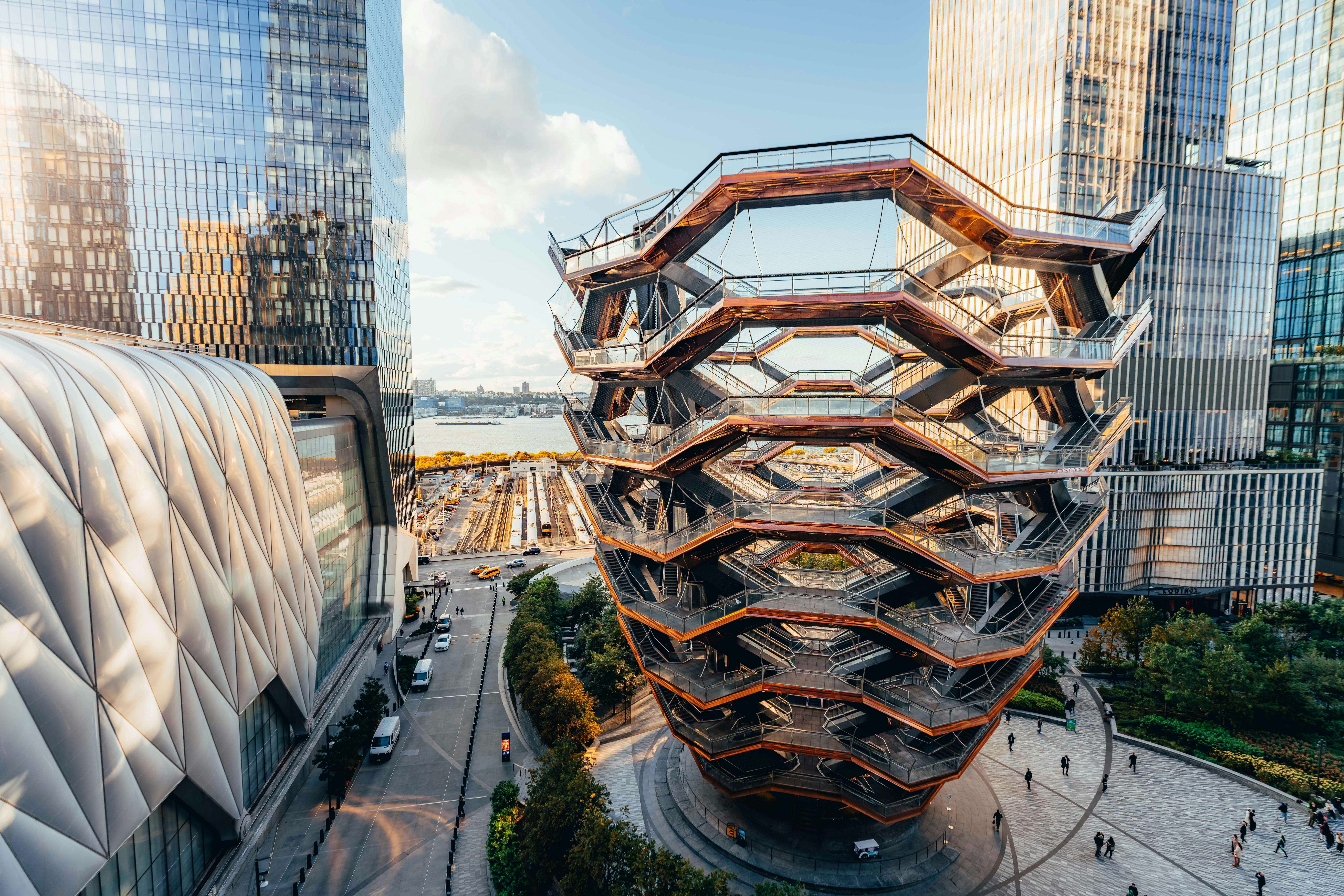Modern, honeycomb-like structure surrounded by tall buildings, overlooking a waterfront and train tracks in an urban setting.
