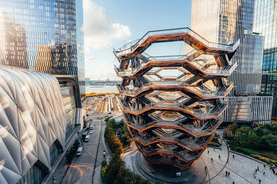 Modern, honeycomb-like structure surrounded by tall buildings, overlooking a waterfront and train tracks in an urban setting.