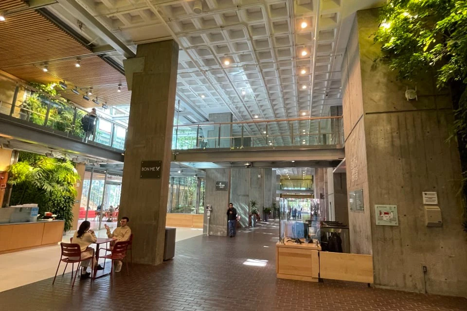 Spacious atrium with high ceiling, featuring modern decor, large windows, and people sitting at tables and walking.