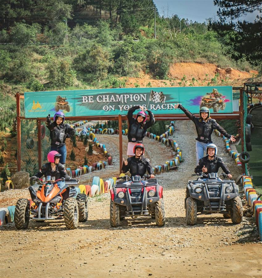 A group of people riding quad bikes at an outdoor track with a banner reading 'Be Champion on Your Racing' in the background.
