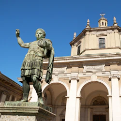 Bronsstaty av en romersk figur framför en historisk byggnad med pelare och välvda fönster, mot en klarblå himmel.