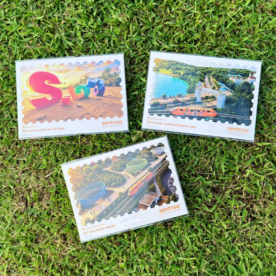 Three Sentosa-themed postcards featuring colorful illustrations, placed on a grass background.