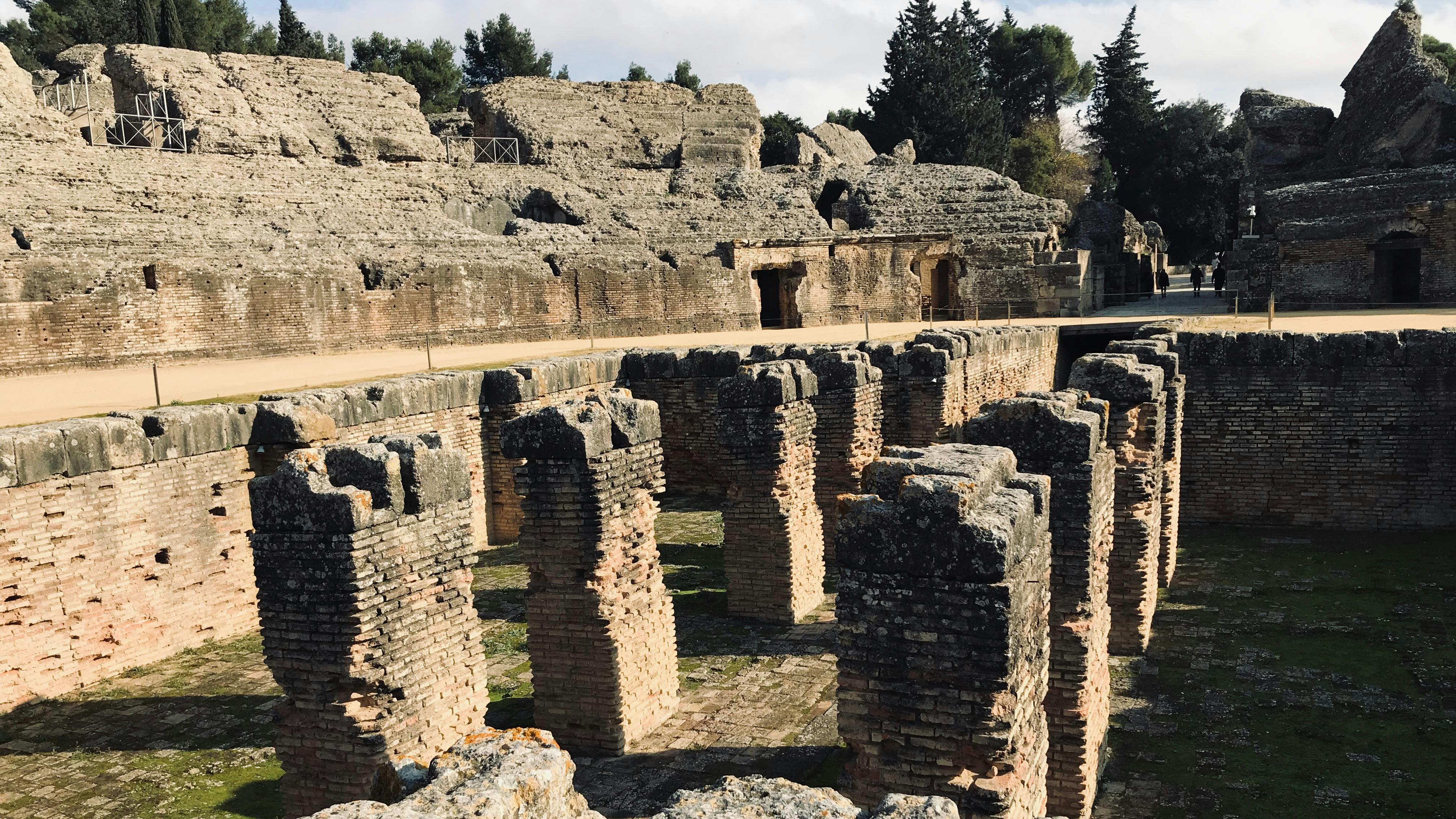 Starobylé kamenné zříceniny s několika cihlovými sloupy na historickém místě, obklopené stromy pod zataženou oblohou.