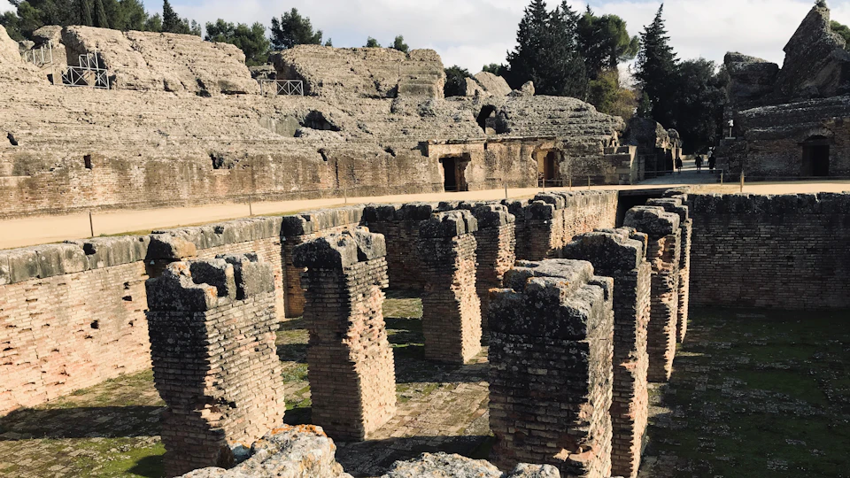 Ancient stone ruins featuring multiple brick columns in a historical site, surrounded by trees under a cloudy sky.