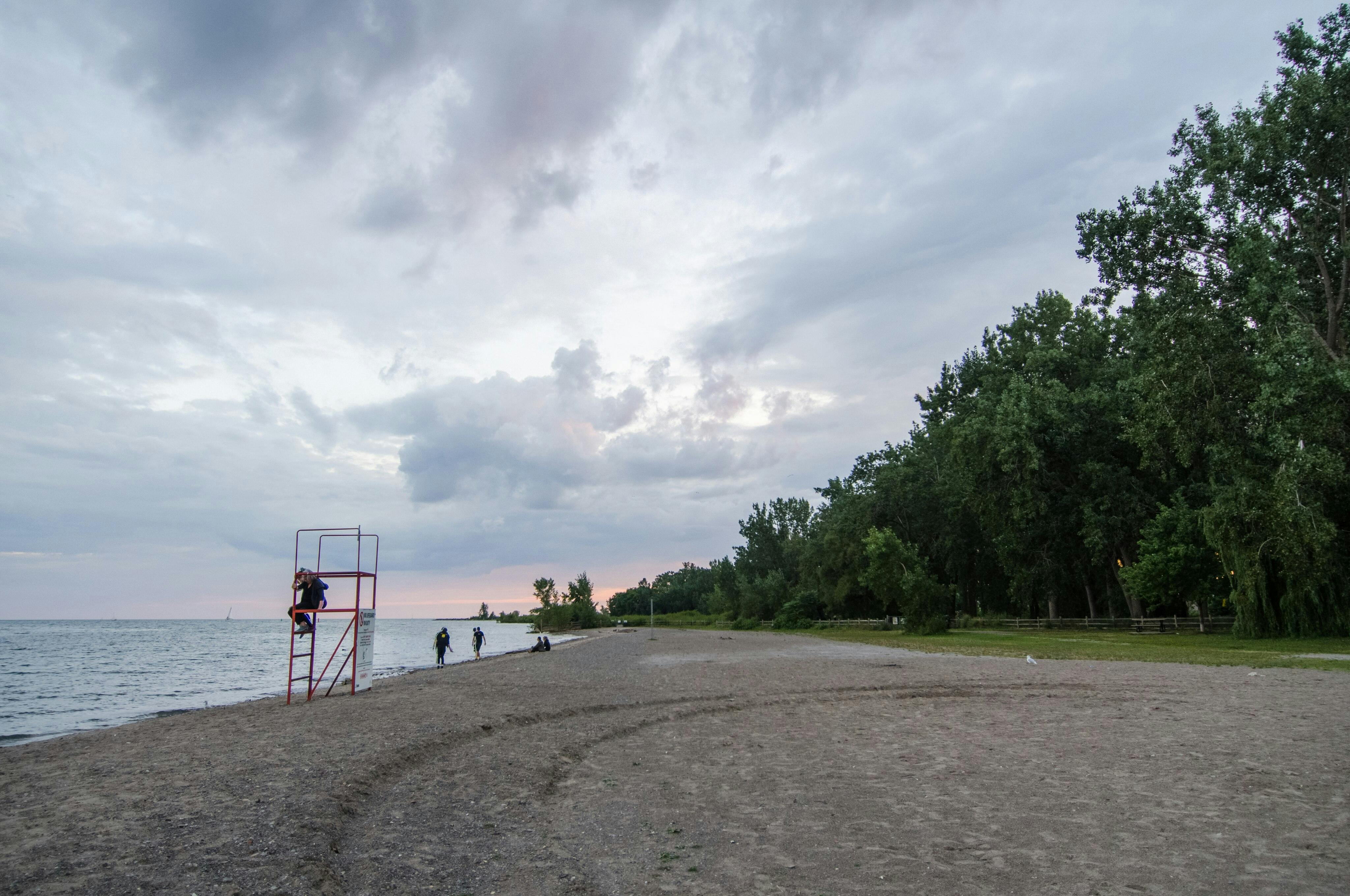 Strand mit bedecktem Himmel, Rettungsschwimmerturm links, Menschen, die am Ufer spazieren gehen, Bäume rechts.