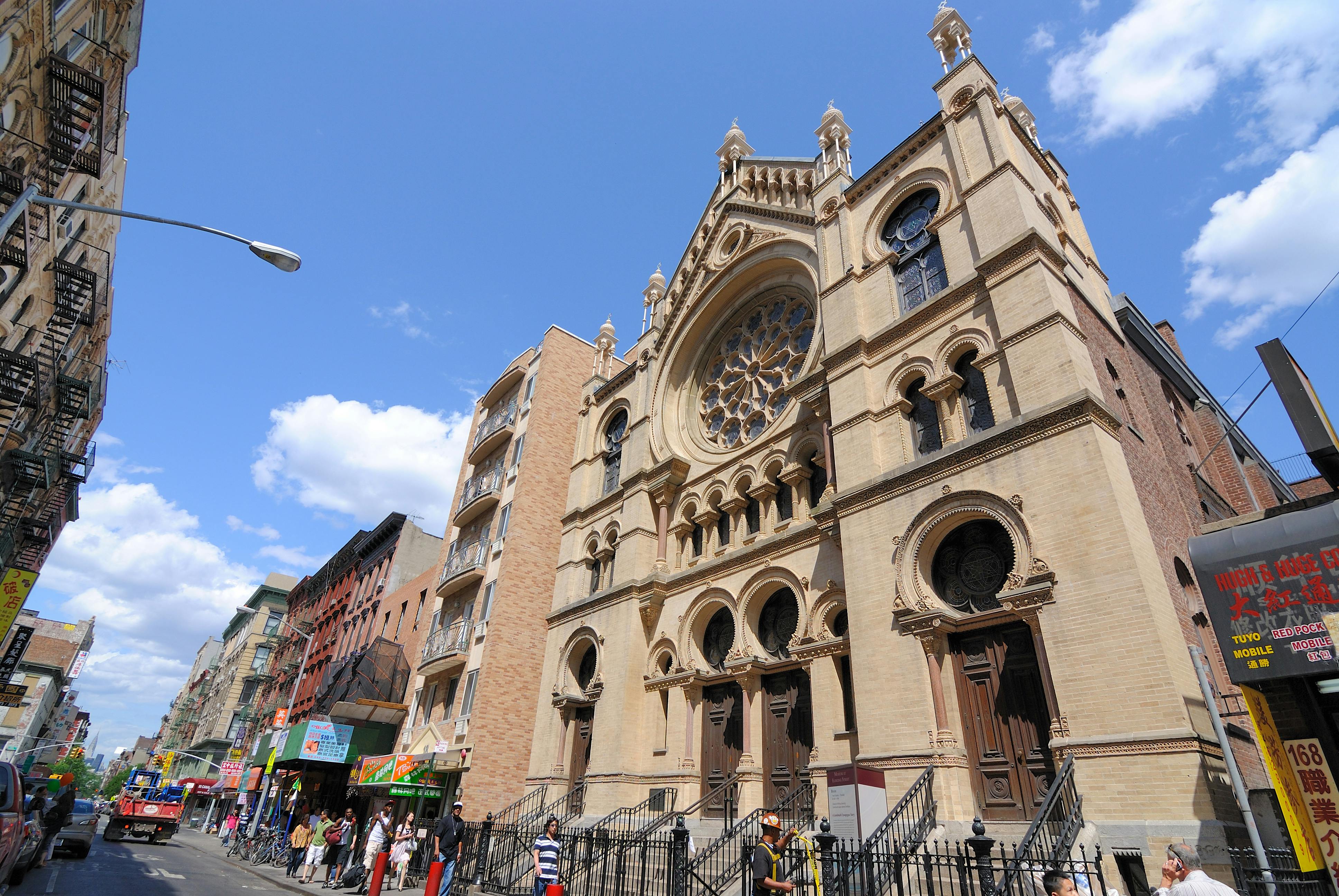 Une grande cathédrale aux pierres ornées et aux portes cintrées se dresse à côté d'une rangée de bâtiments. Des gens marchent sur le trottoir.
