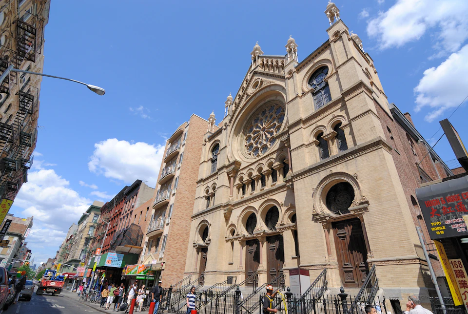 A large cathedral with ornate stonework and arched doors stands adjacent to a row of buildings. People are walking on the sidewalk.