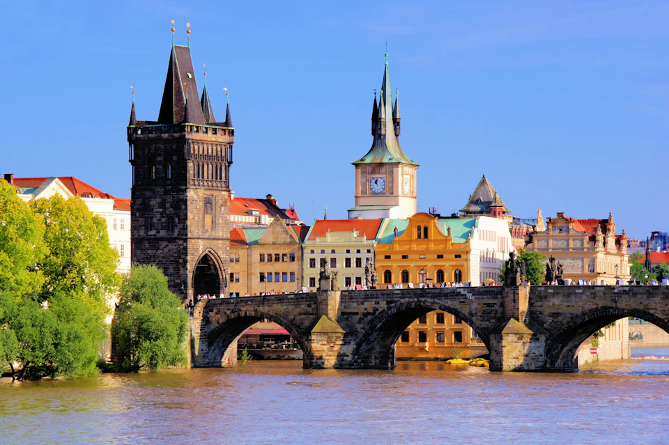 A stone bridge over a river with historical buildings and towers in the background under a clear blue sky.