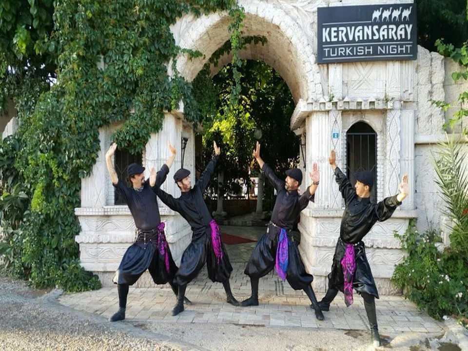 Four men in black traditional attire with purple sashes perform a synchronized dance outside a decorated stone archway.