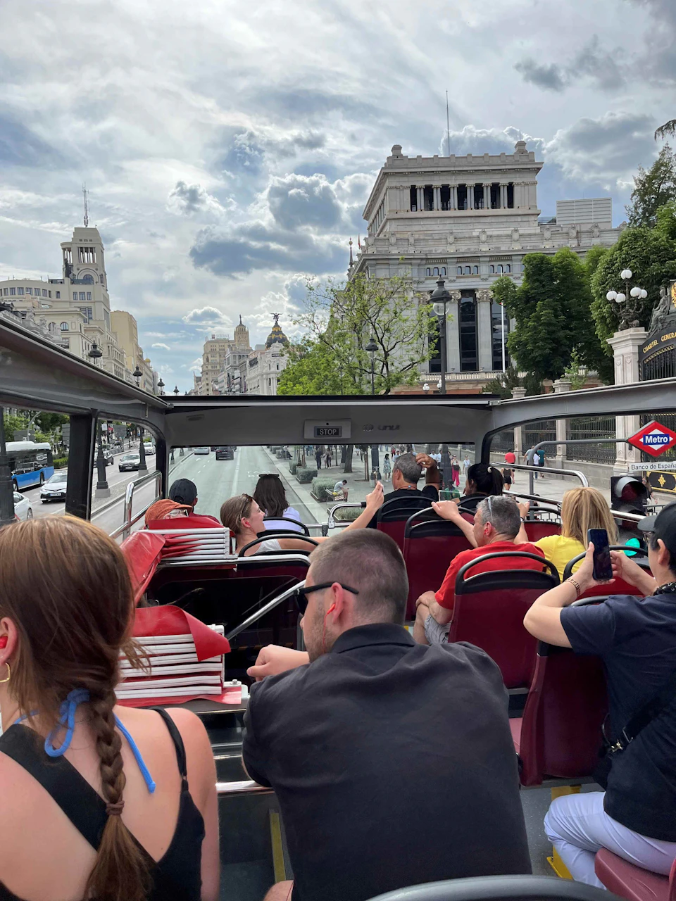 People seated on the upper deck of a double-decker tour bus riding through a city with historic buildings in the background.