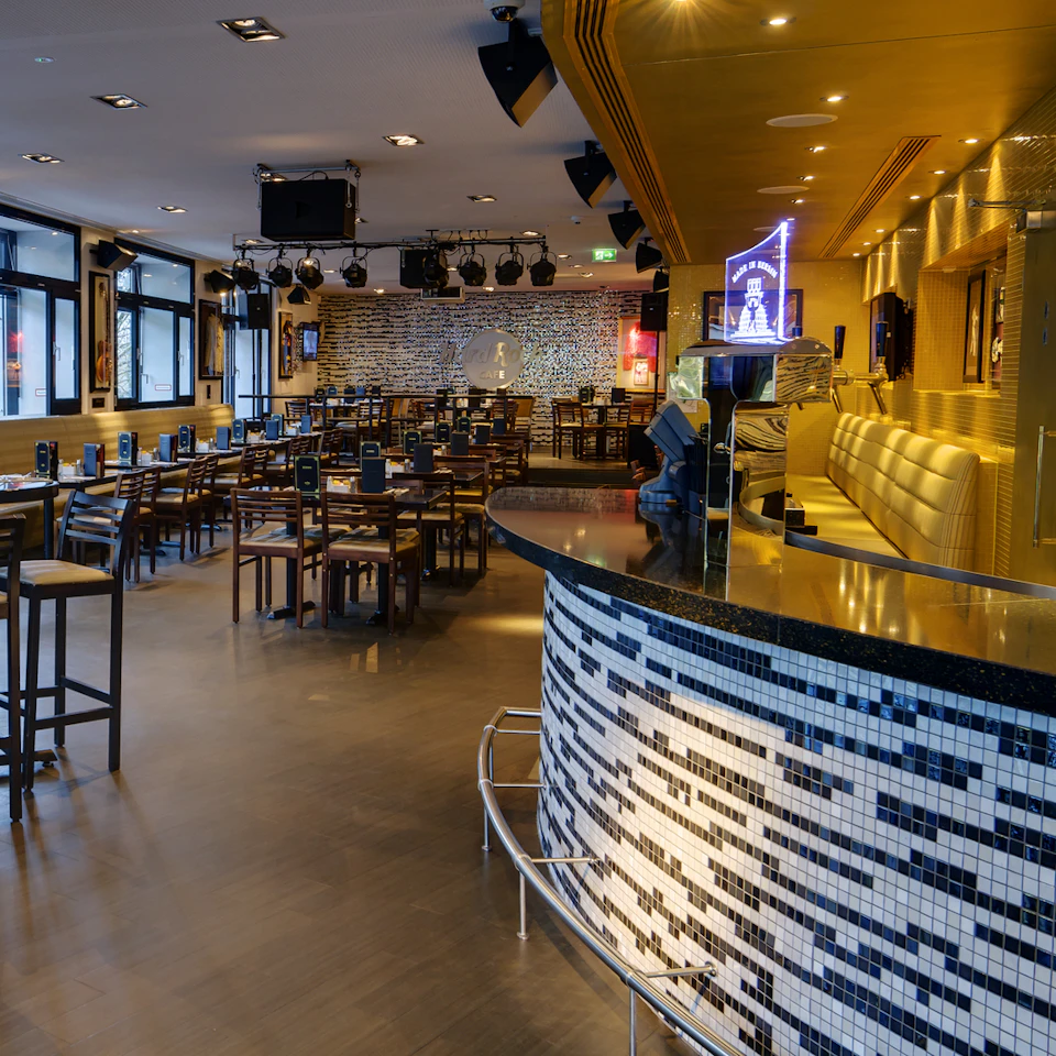 A modern bar with a tiled counter, high tables and chairs, yellow booths, and a stage with a drum set under spotlights.