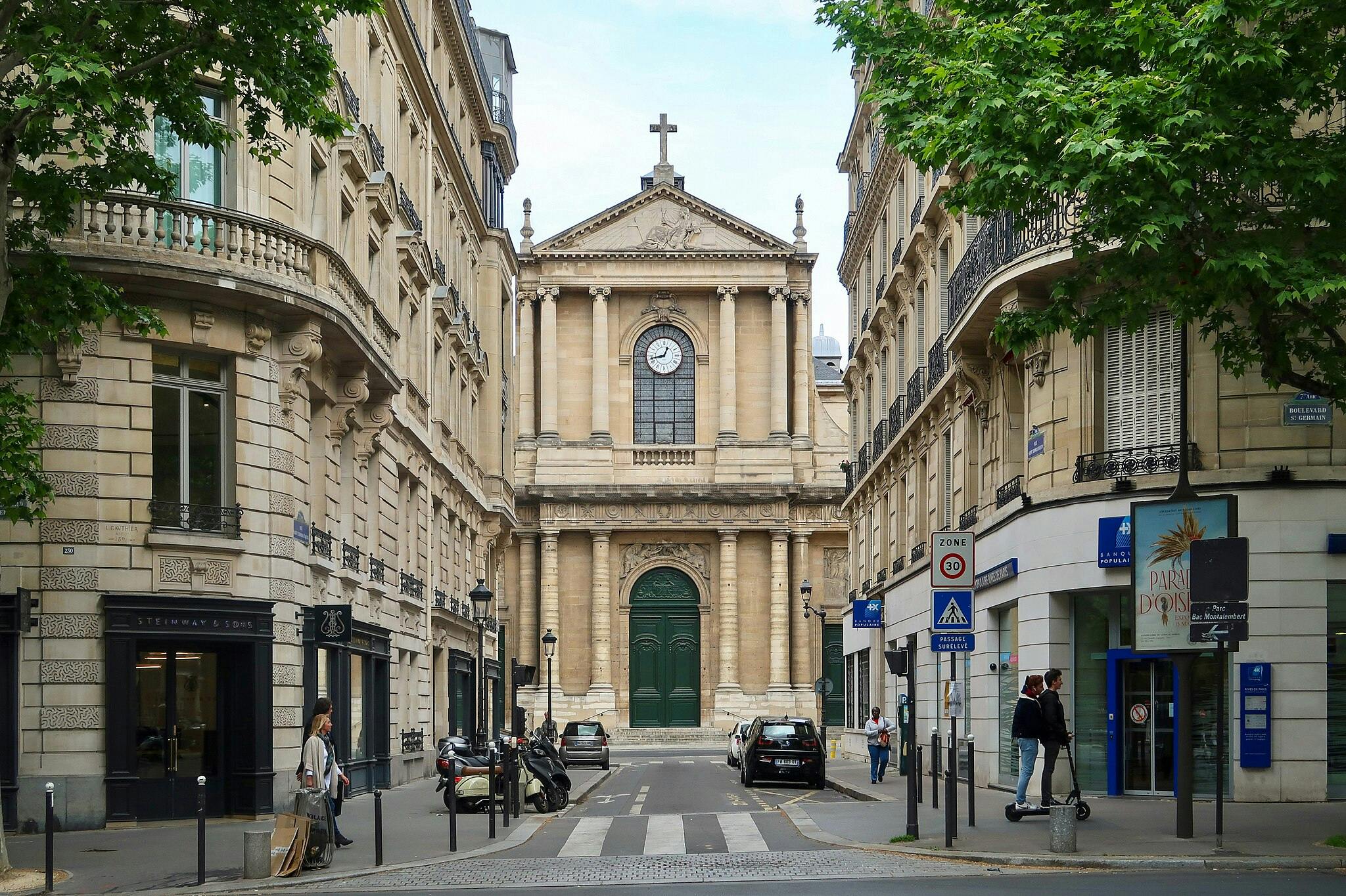 Urban street scene with classic European architecture, a church with a clock, parked vehicles, pedestrians, and a cyclist.