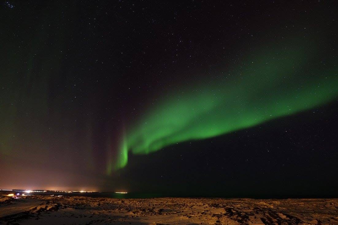 Zelená polární záře borealis prolétá hvězdnou noční oblohou nad zasněženou krajinou se světly viditelnými na obzoru.