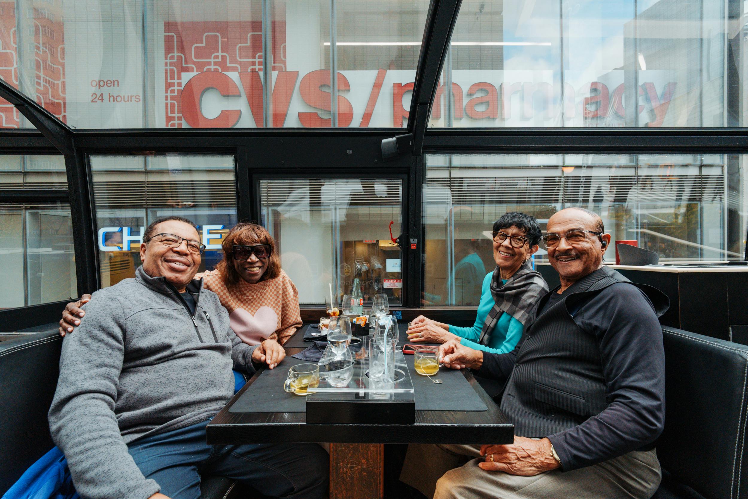 Trois personnes sourient alors qu'elles sont assises à une table avec des boissons dans un espace vitré. Une enseigne de pharmacie est visible à travers la fenêtre.