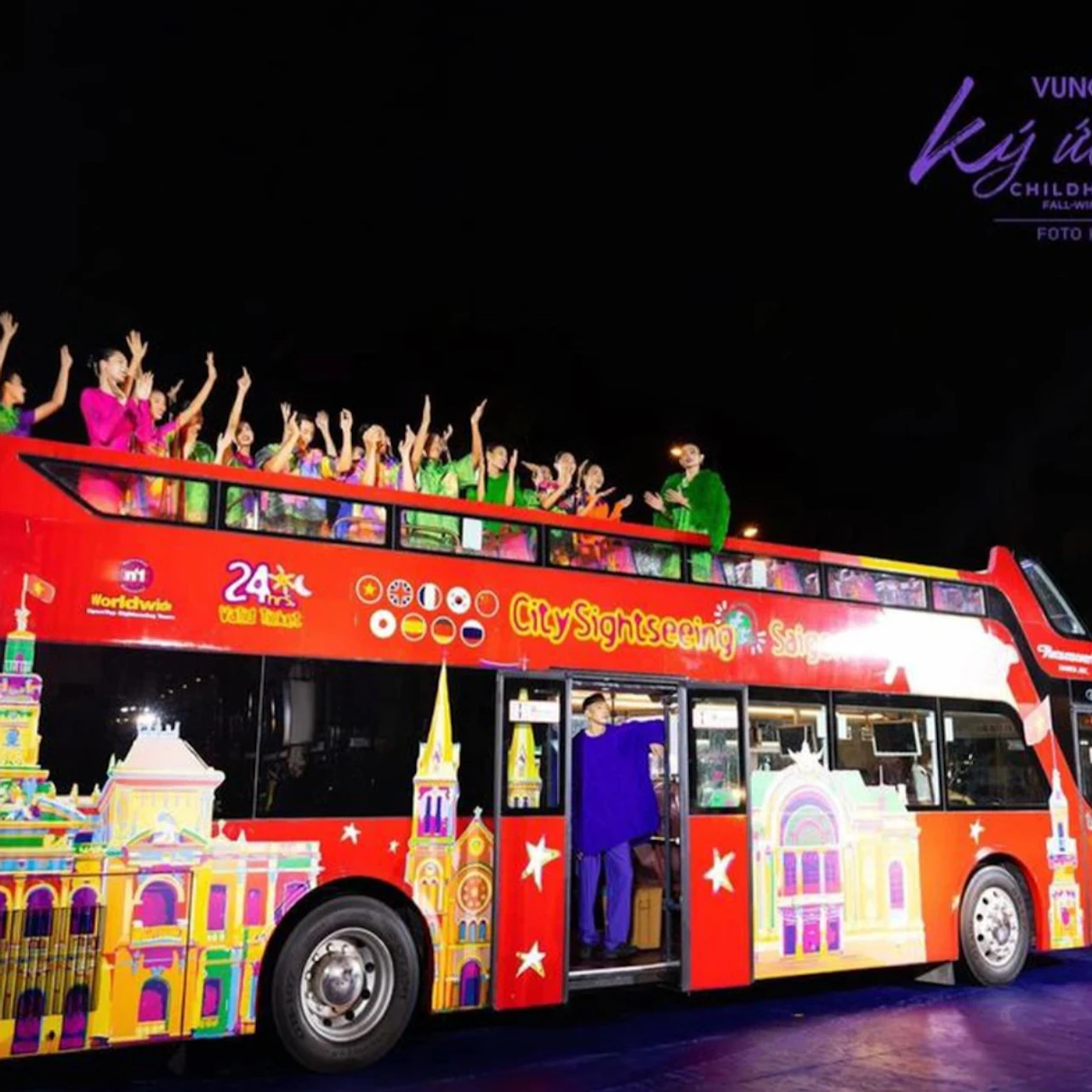 A brightly colored double-decker sightseeing bus with passengers on the open upper deck waving excitedly at night.