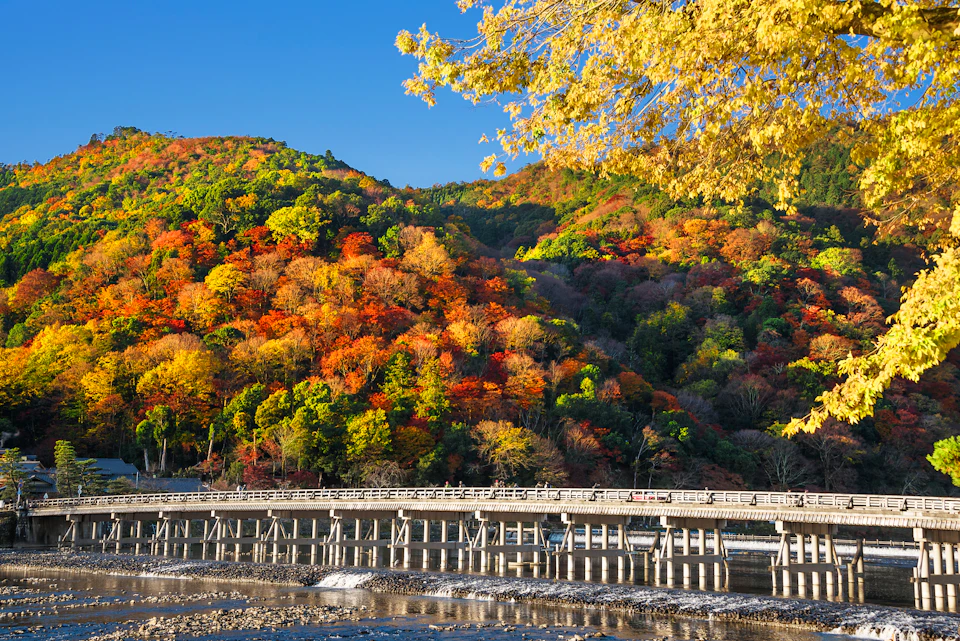 A scenic view of a wooden bridge over a river, with colorful autumn foliage on hills under a clear blue sky.