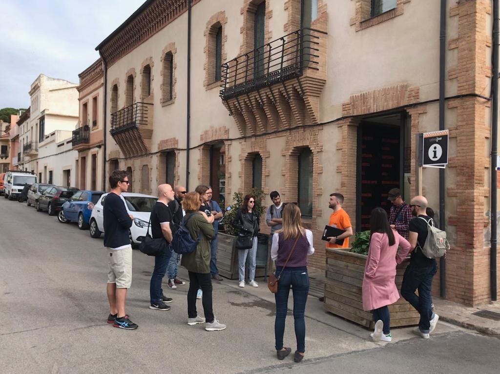 A group of people standing on a street in front of a building with arched windows and balconies, engaging in conversation.