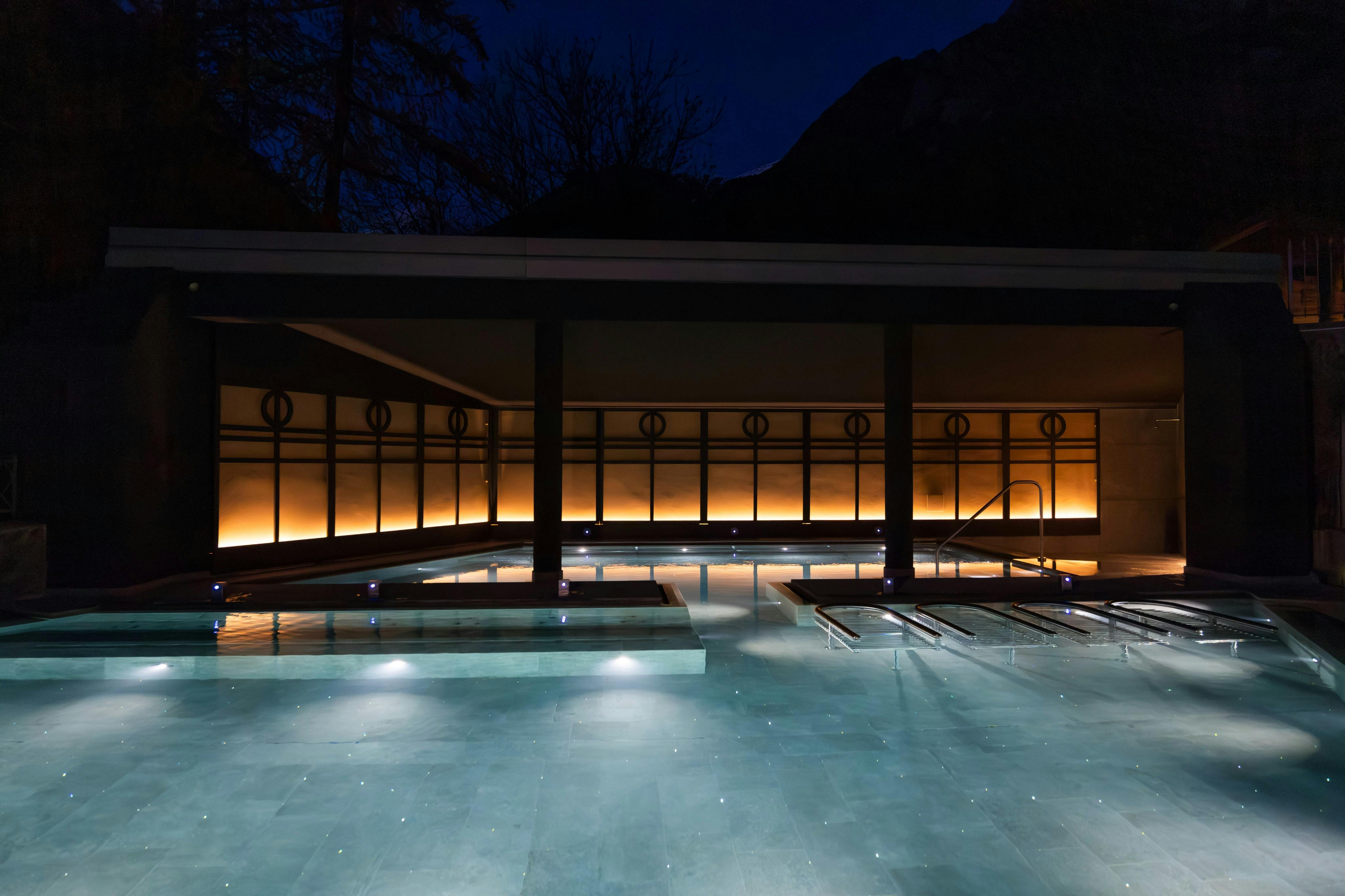 A modern pool at night with illuminated water and a lit, minimalist architecture in the background.