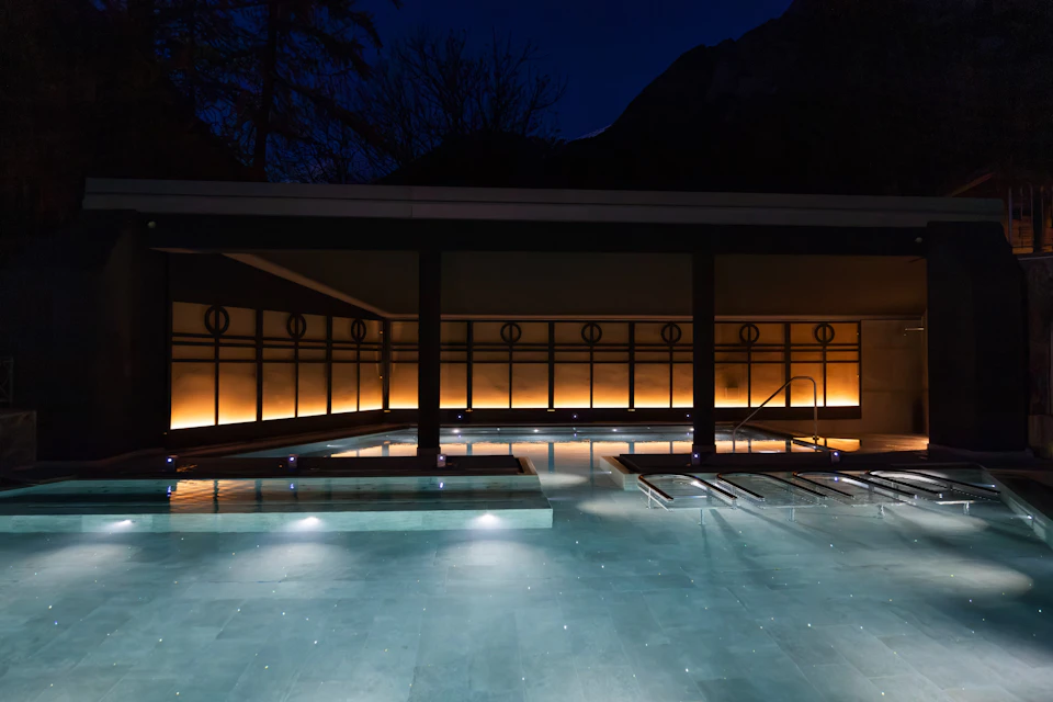 A modern pool at night with illuminated water and a lit, minimalist architecture in the background.
