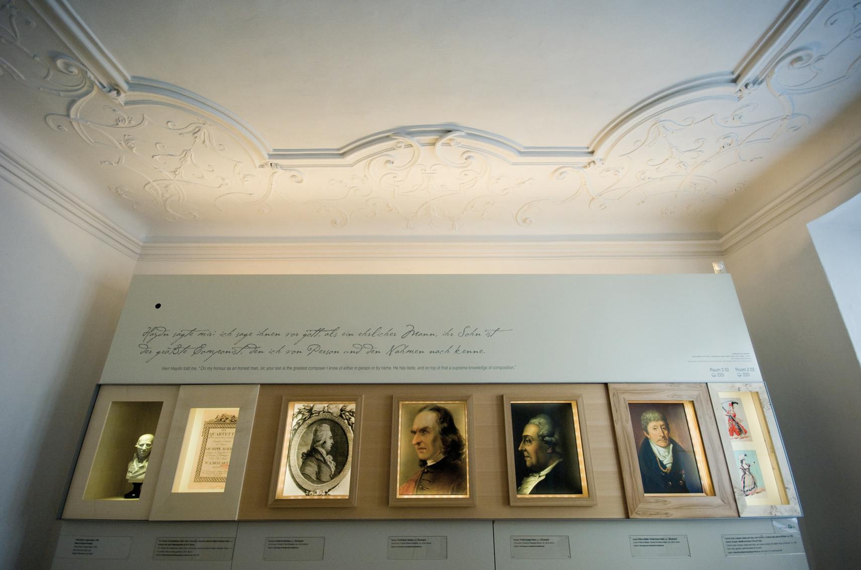 Kunstwerken en portretten tentoongesteld in een museum onder een sierlijk plafond met decoratieve tekst boven de tentoongestelde stukken.