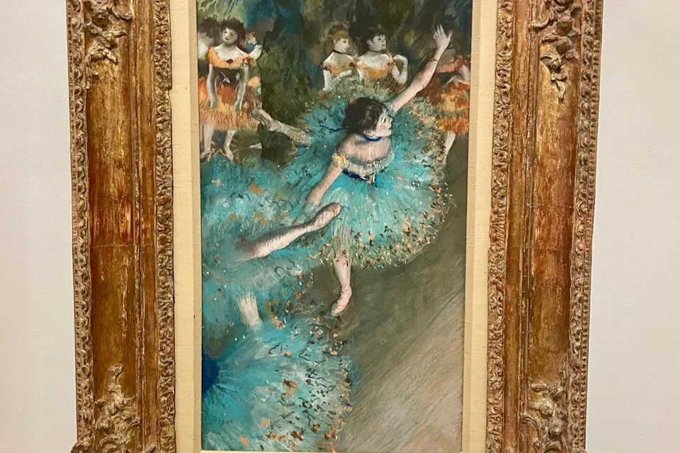 A framed painting depicting ballerinas dressed in blue tutus performing against a backdrop of green foliage.