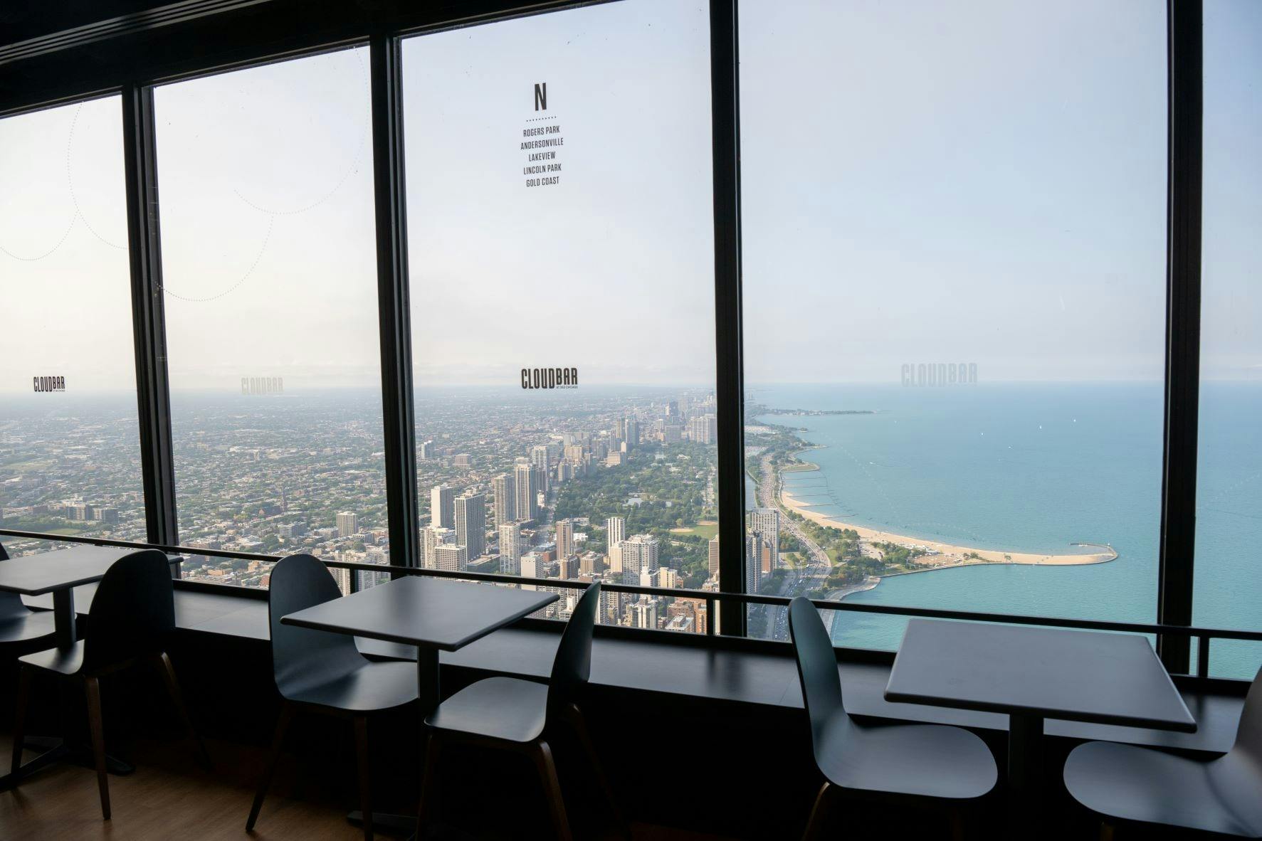 Tables and chairs by large windows overlooking a cityscape and a coastline with buildings extending along a sandy beach.